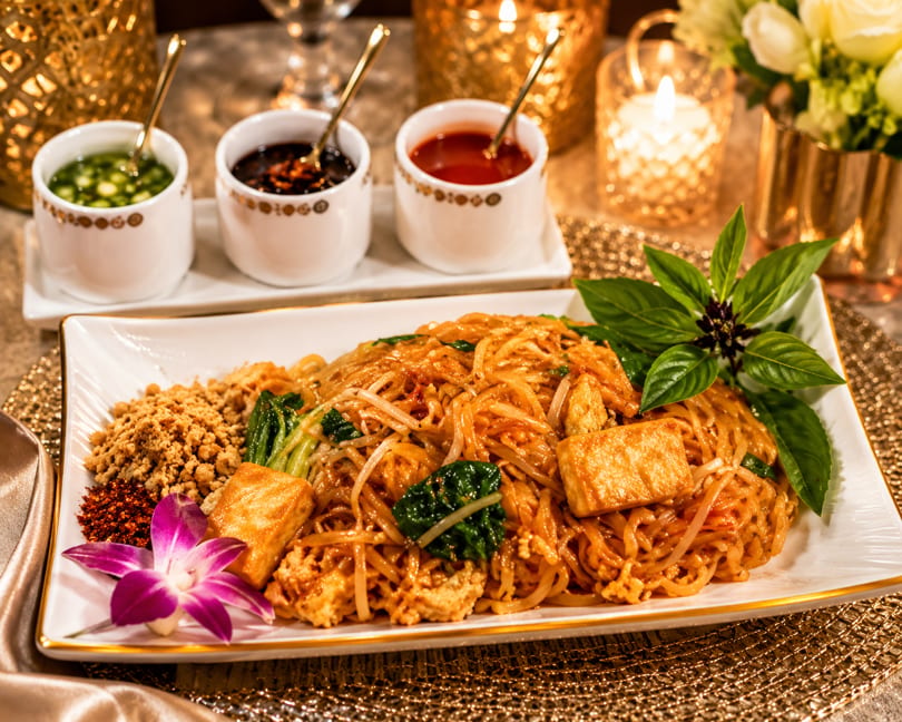 Vibrant Pad Thai noodles with tofu, peanuts, chili, basil, and an orchid, served with sauces in a warm, candlelit setting.