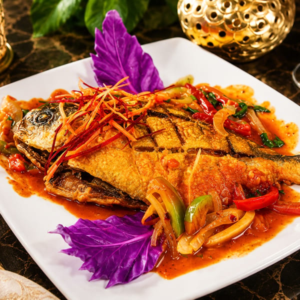 Golden fried fish with vibrant red sauce, bell peppers, and shredded garnish on a white plate.