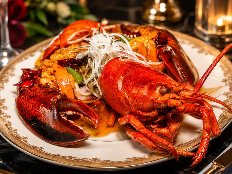A whole red lobster served with vegetables and sauce on a decorative plate.