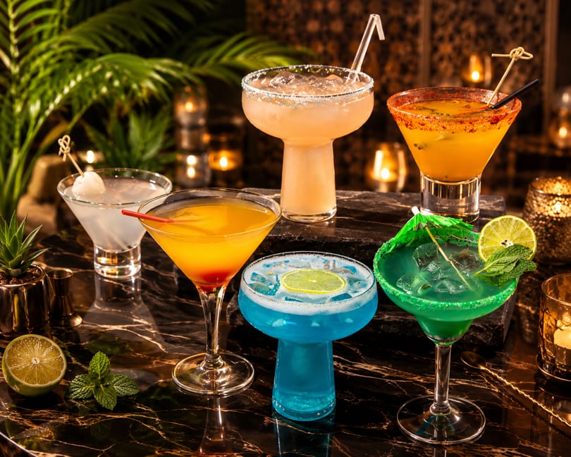 Six vibrant, garnished cocktails on a dark marble bar with tropical plants and warm lighting.