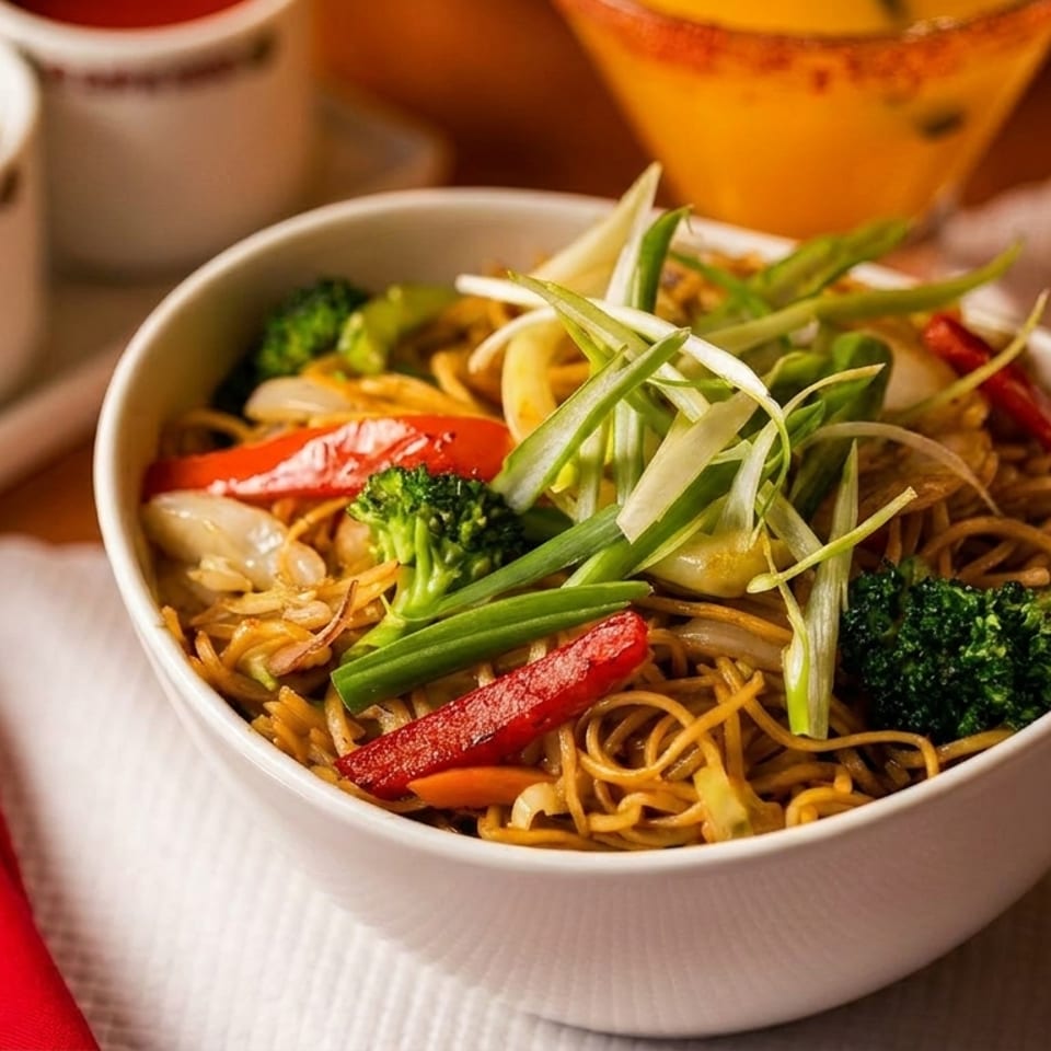 A white bowl of lo mein noodles with broccoli, red peppers, and green onions.