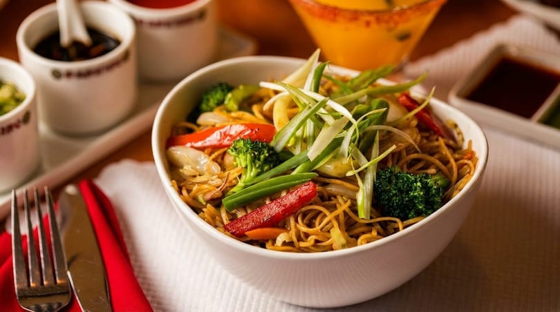 A white bowl of lo mein noodles with broccoli, red peppers, and green onions.