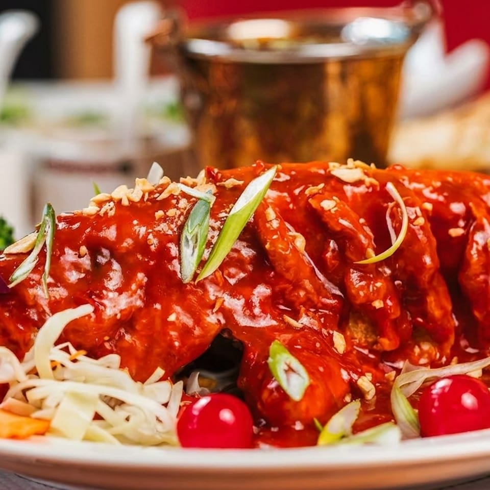 Whole fried fish covered in red sauce, garnished with vegetables, cherries, and orchids.