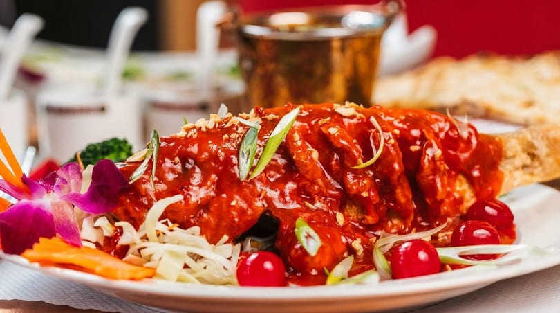 Whole fried fish covered in red sauce, garnished with vegetables, cherries, and orchids.