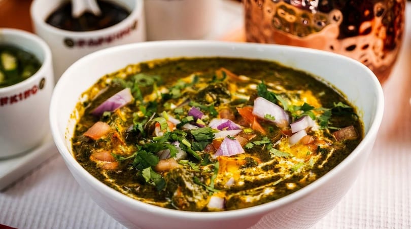 A white bowl filled with green curry, topped with chopped red onions, tomatoes, and cilantro.