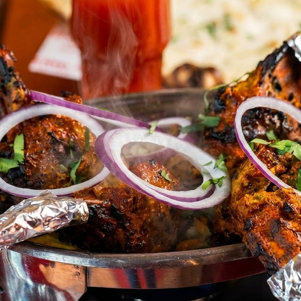 Steaming hot tandoori chicken drumsticks with red onion rings and cilantro in a metal serving dish.