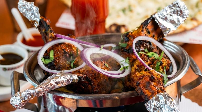 Steaming hot tandoori chicken drumsticks with red onion rings and cilantro in a metal serving dish.