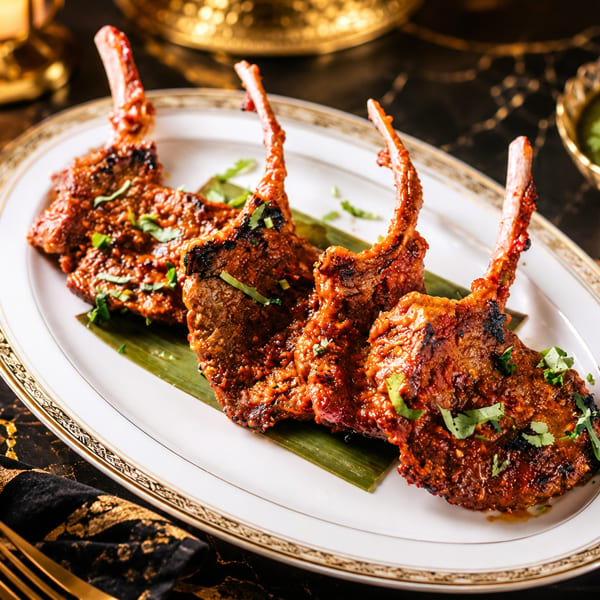 Four spiced lamb chops on a banana leaf, served on a gold-trimmed plate with green sauce.