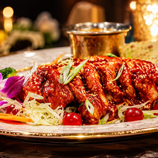 Glazed chicken with sesame seeds, green onions, fresh vegetables, flatbread, and a metal bowl on a decorative plate.